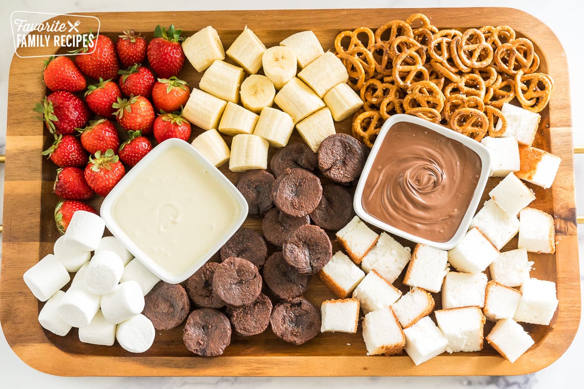 A charcuterie board with white chocolate, and milk chocolate and strawberries, pretzels, marshmallows, bananas, pound cake, and brownie pieces.