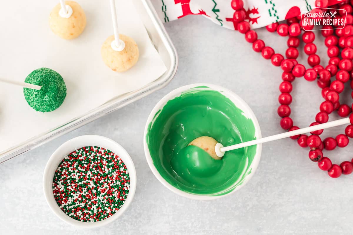 Cake Pop being dipped into a bowl of melted green candy melts. Sprinkles on the side.