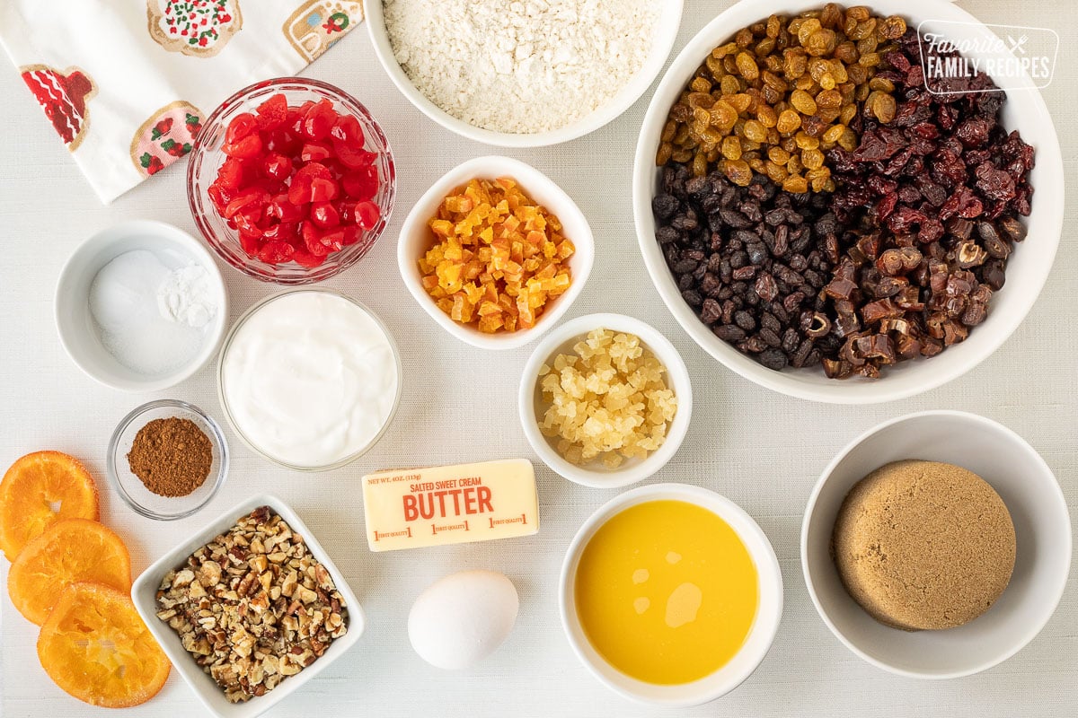 Ingredients to make Fruit Cake including golden raisins, raisins, dates, dried cherries, brown sugar, orange juice, flour, sweetened dried orange, candied ginger, egg, butter, sour cream, chopped nuts, cherries, pumpkin pie spice, salt, baking powder and baking soda.