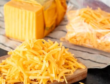 Grated cheddar cheese on a cutting board next to a block of cheese and bag of freeze cheese.