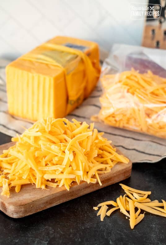 Grated cheddar cheese on a cutting board next to a block of cheese and bag of freeze cheese.