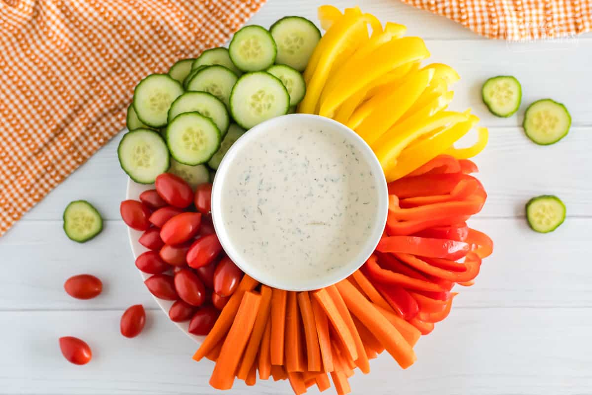 A white plate of veggies with a bowl full of ranch dressing. 