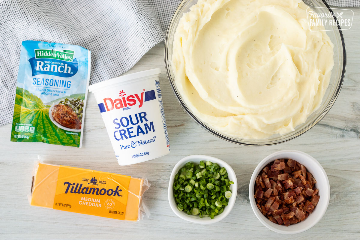 Ingredients for Loaded Mashed Potatoes including mashed potatoes, sour cream, ranch dressing seasoning, cheddar cheese, green onions and bacon.