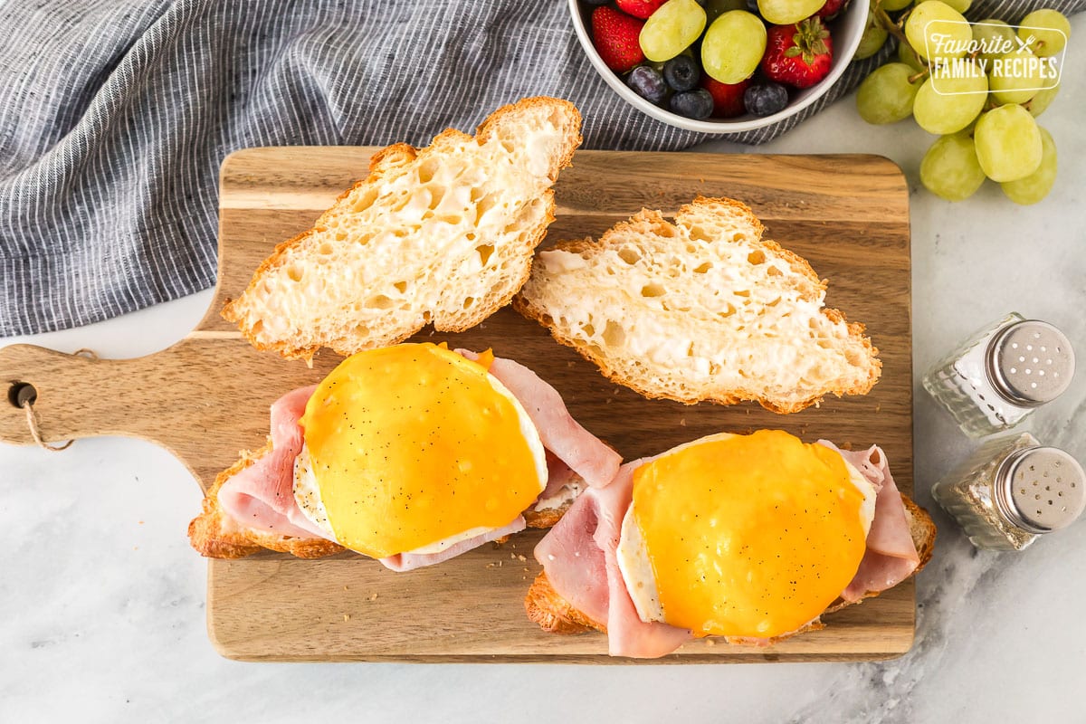 Two croissants on a cutting board topped with ham, egg, cheese, salt and pepper with fresh fruit on the side.