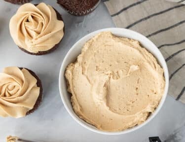 Bowl of Peanut Butter Frosting with a piping bag of Peanut Butter Frosting and chocolate cupcakes.