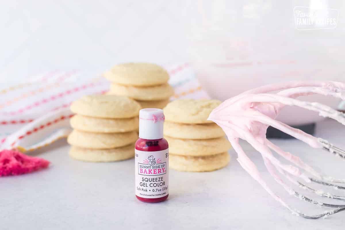 Pink gel color bottle next to whisk of pink frosting and cookies in the background.