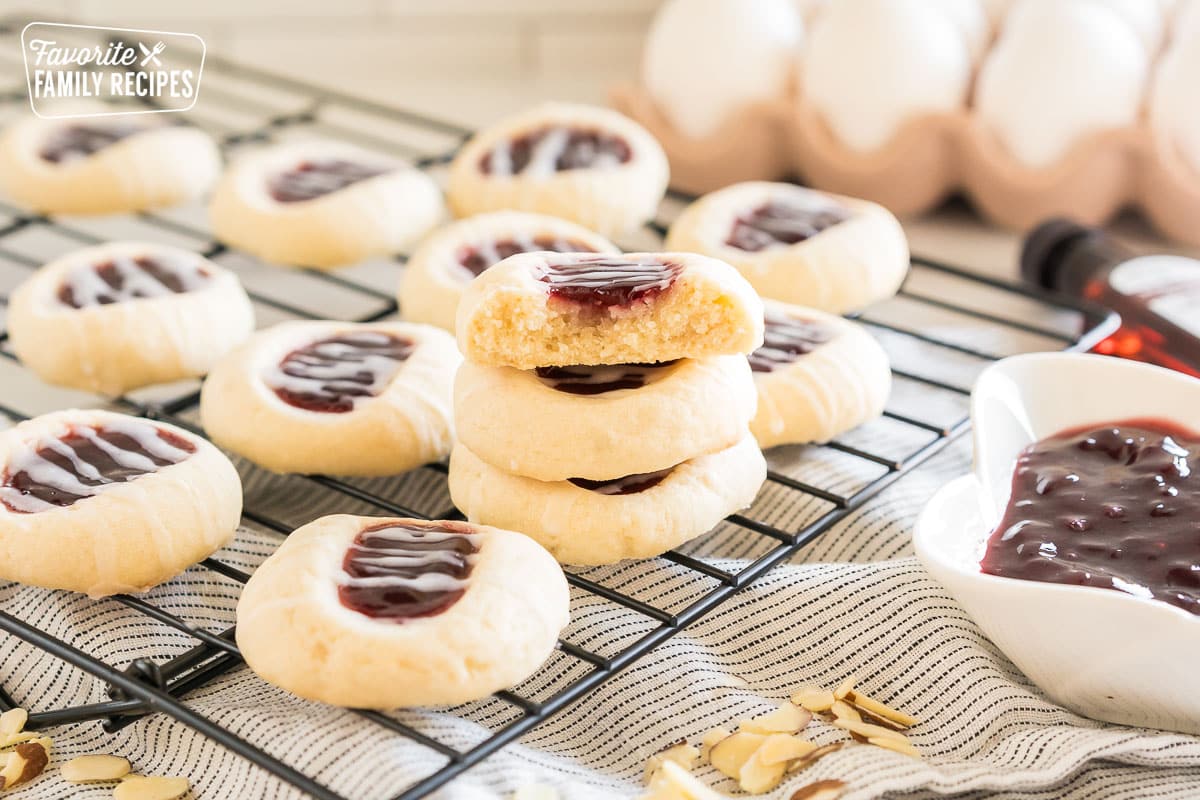 a stack of Raspberry Thumbprint cookies on a wire rack with one broken in half so you can see the inside