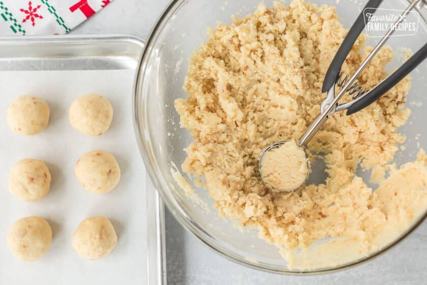 Mixing bowl with cake pop mixture and cookie scoop. Cake pop balls placed to the side on a baking sheet.
