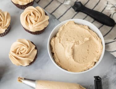 Bowl of Peanut Butter frosting next to decorated cupcakes and piping bag.