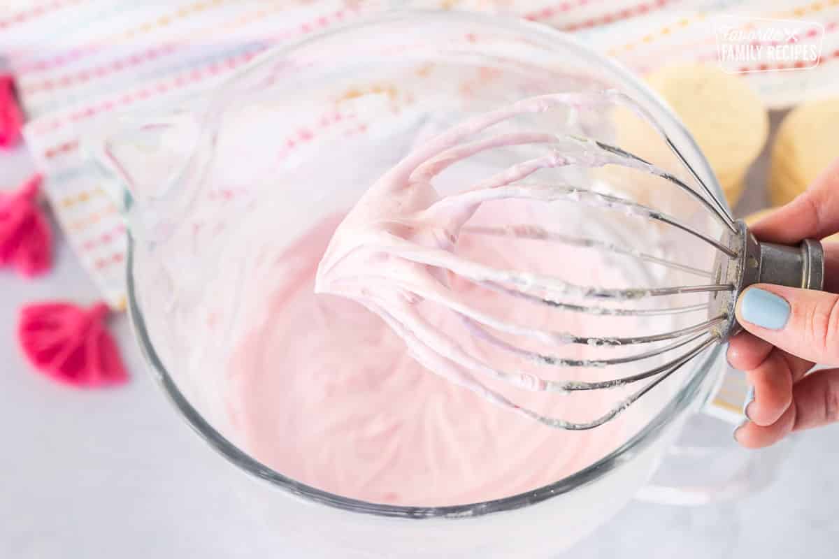 Hand holding a whisk of pink sugar cookie frosting.
