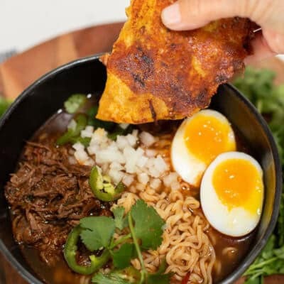 A birria taco being dipped into a bowl of ramen