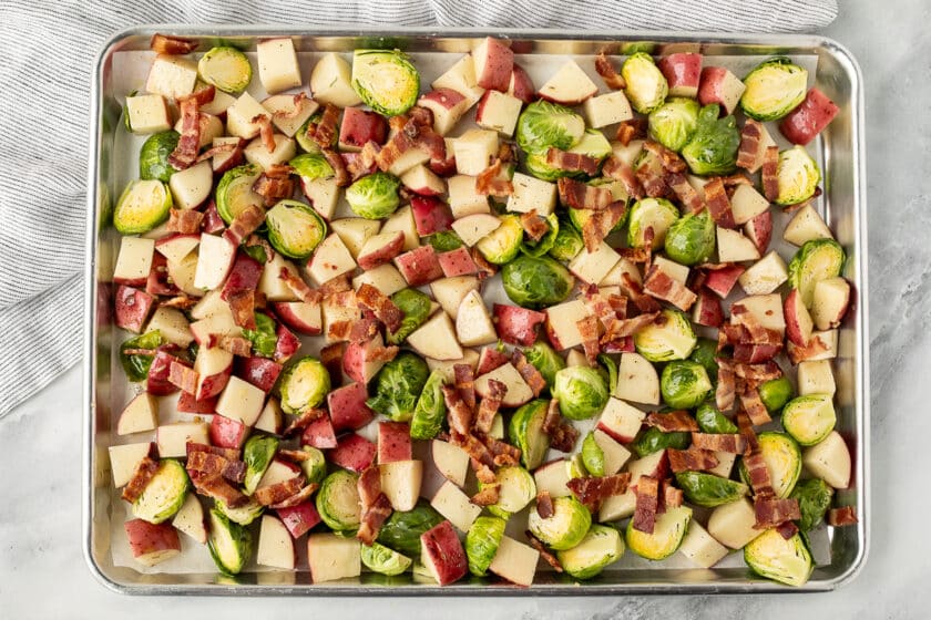 Baking sheet with cut red potatoes an cut Brussels sprouts topped with bacon pieces.
