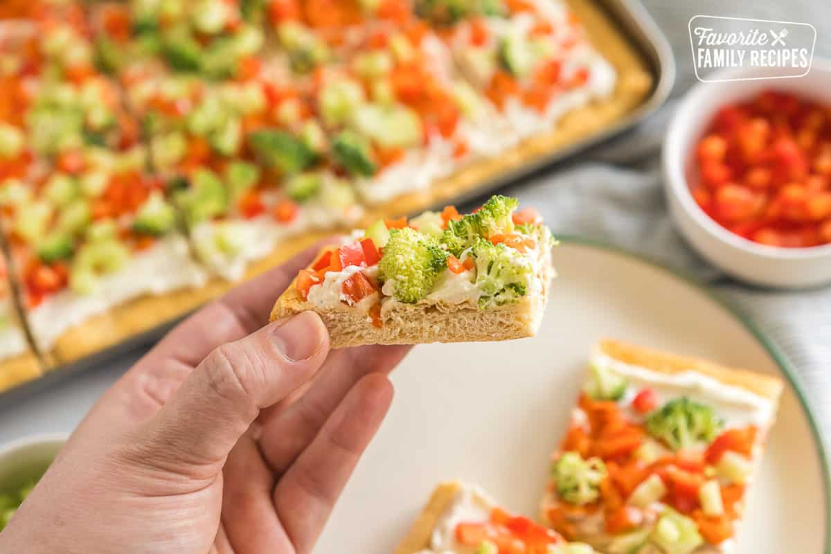 A hand holding a square of crescent roll veggie pizza