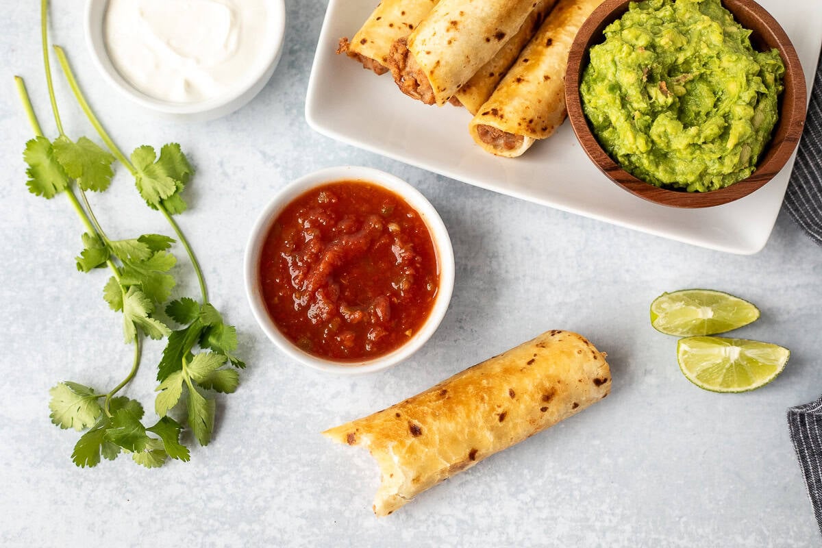 Bite missing from a Crisp Bean Burritos. Plate of Crisp Bean Burritos on the side with a bowl of guacamole. Salsa bowl, lime slices and a bowl of sour cream on the side.