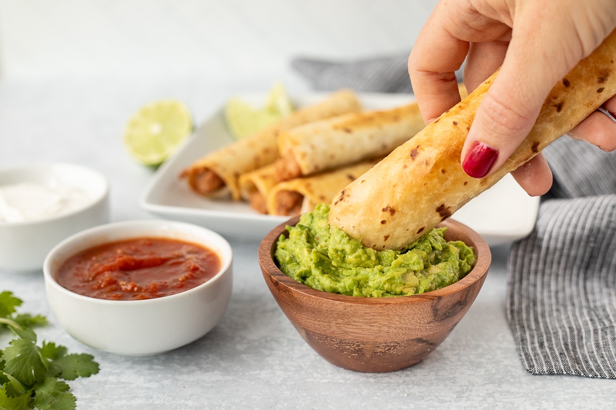 Dipping a Crisp Bean Burrito into a bowl of guacamole. Salsa and sour cream bowls on the side.