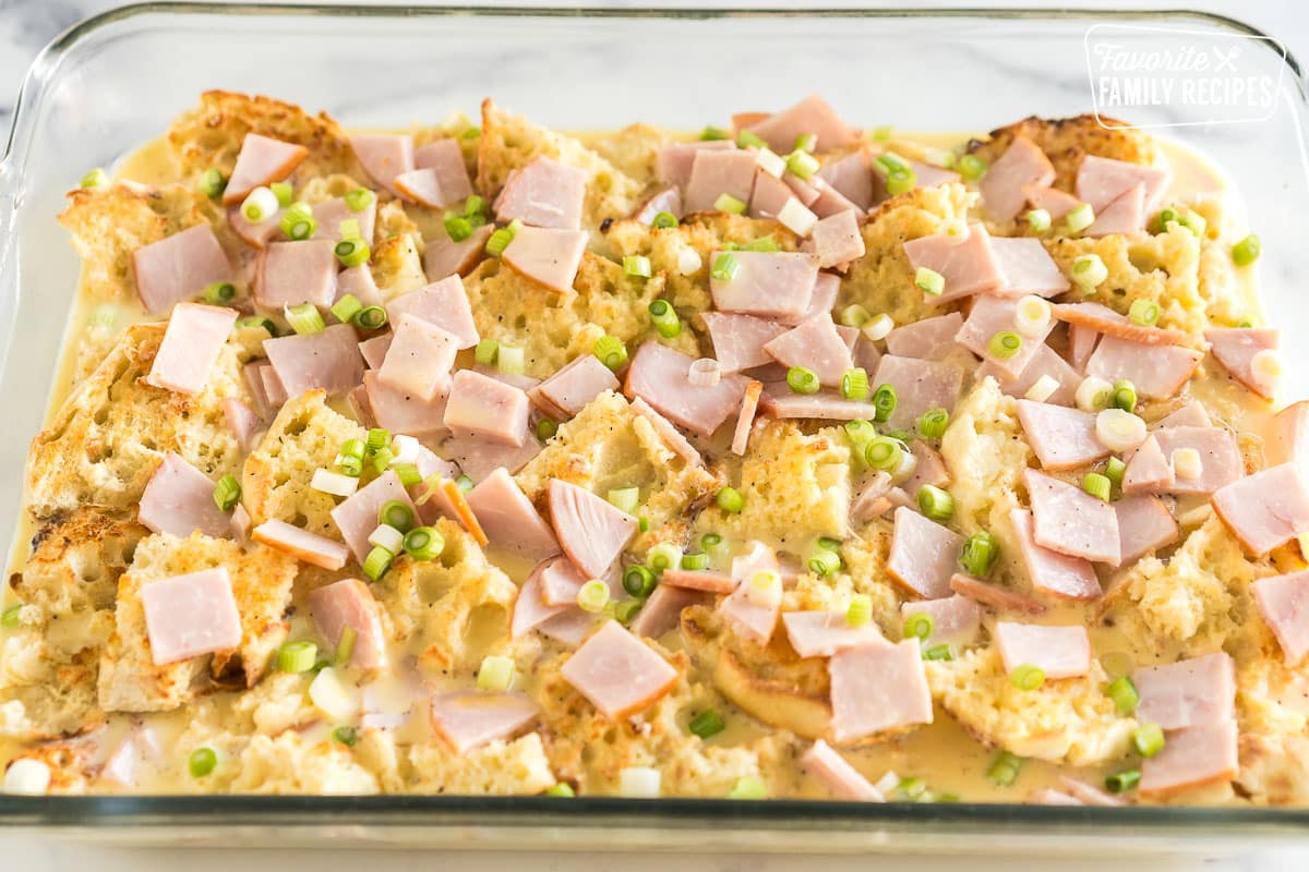 toasted and buttered english muffins topped with sliced canadian bacon and green onions and covered with an egg mixture in a baking dish