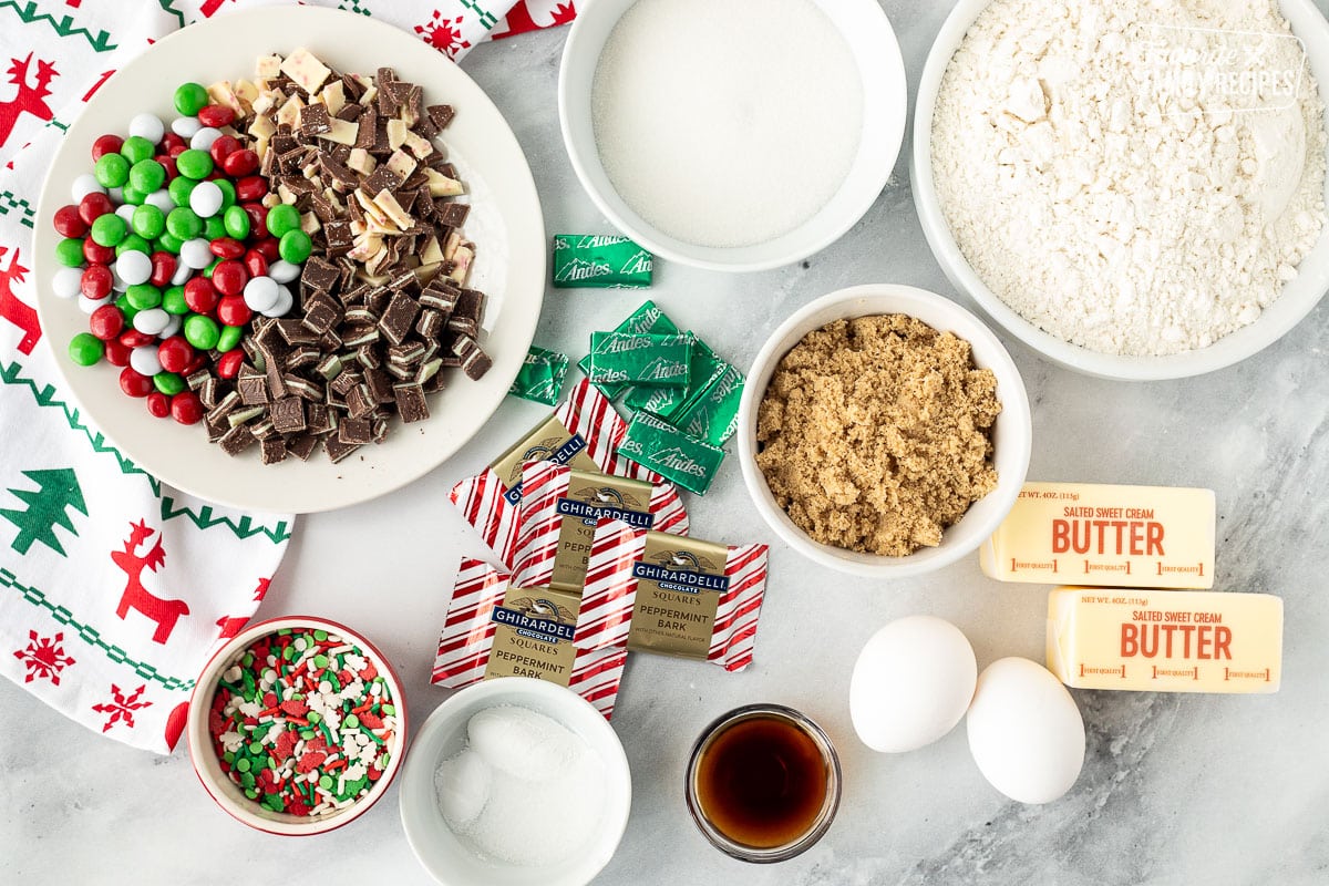 Ingredients to make Elf Cookies including flour, sugar, brown sugar, butter, eggs, vanilla, sprinkles, salt, baking soda, baking powder, andes mints, peppermint bark and chocolate candies.