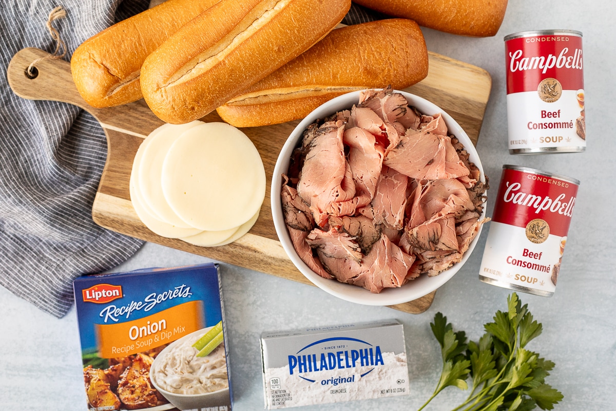Ingredients to make French Dip Sandwiches including sliced roast beef, two cans of beef consommé, cream cheese, onion soup mix, provolone cheese and French rolls.