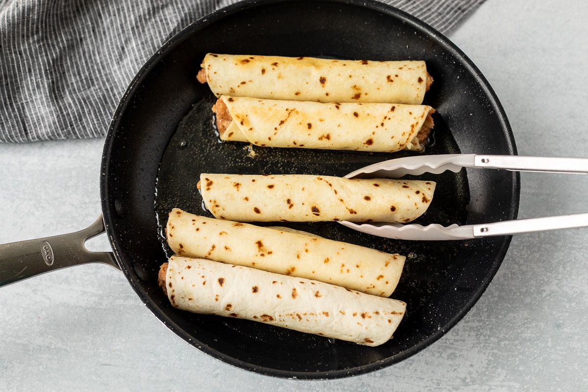 Frying flour tortilla burritos in a skillet with oil.