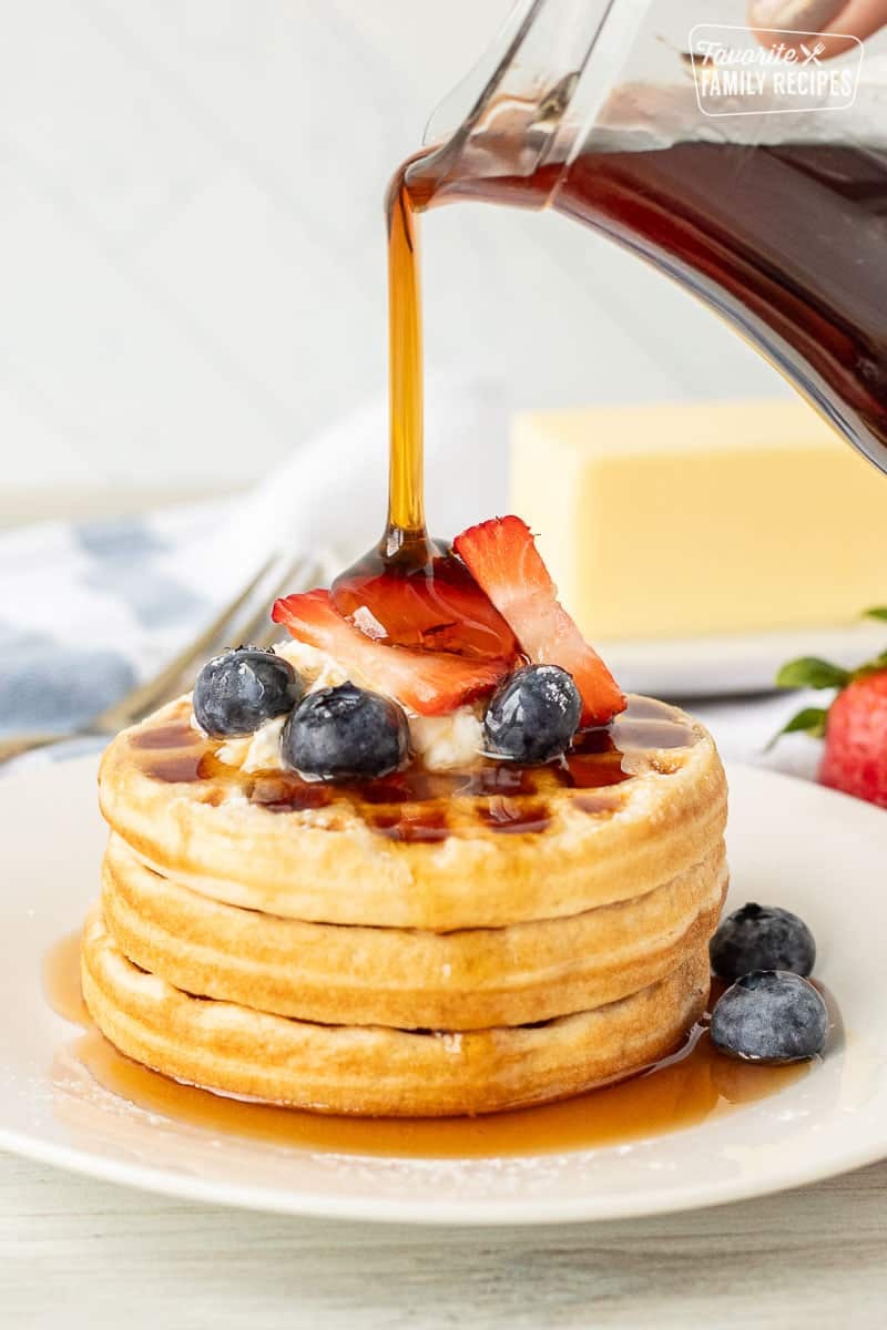 Jar pouring homemade maple syrup over waffles topped with cream and berries.