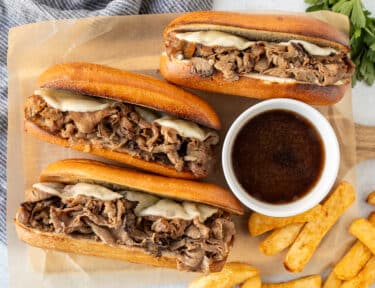 Three French Dip Sandwiches with toasted French bread, sliced roast beef and melted provolone cheese. Bowl of au jus and French fries on the side.