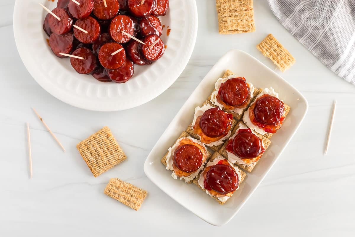 A plate with crackers topped with cream cheese, jalapeno jelly and a kielbasa round. More kielbasa in the Background as well.