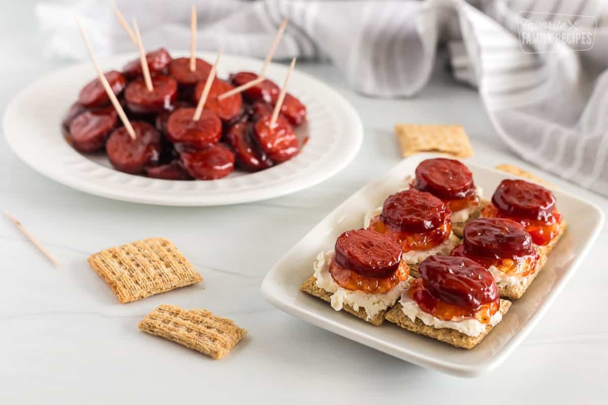 A plate with crackers topped with cream cheese, jalapeno jelly and a kielbasa round. More kielbasa in the Background as well.