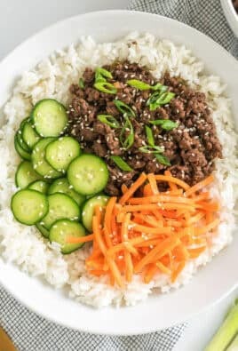 A bowl of rice with ground meat, sliced cucumbers, shredded carrots, and green onions