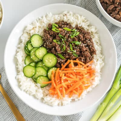 A bowl of rice with ground meat, sliced cucumbers, shredded carrots, and green onions