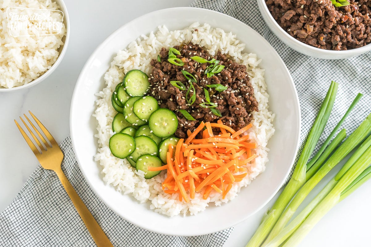 A bowl of rice with ground meat, sliced cucumbers, shredded carrots, and green onions.