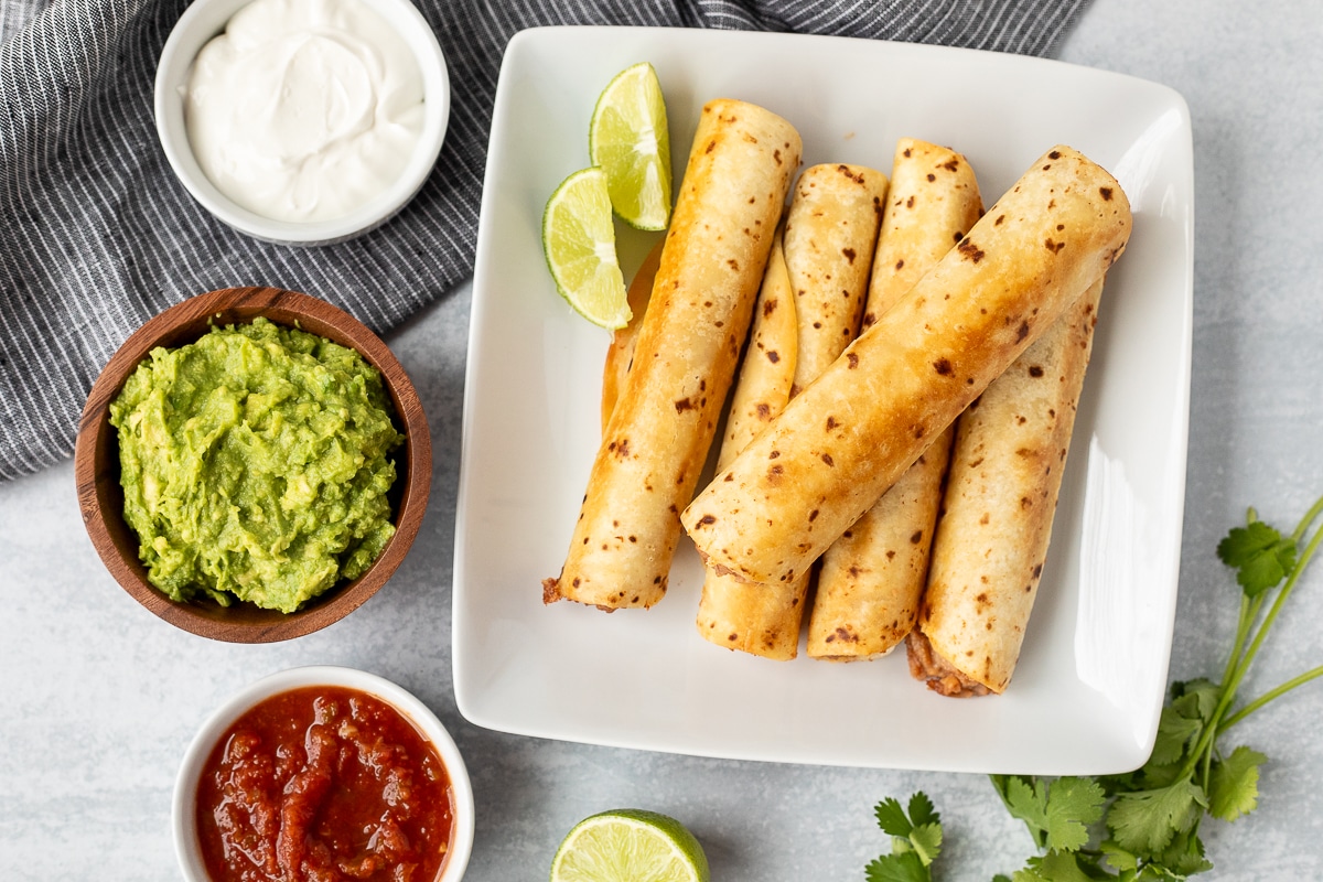 Five Crisp Bean Burritos on a plate. Guacamole, sour cream and salsa on the side.