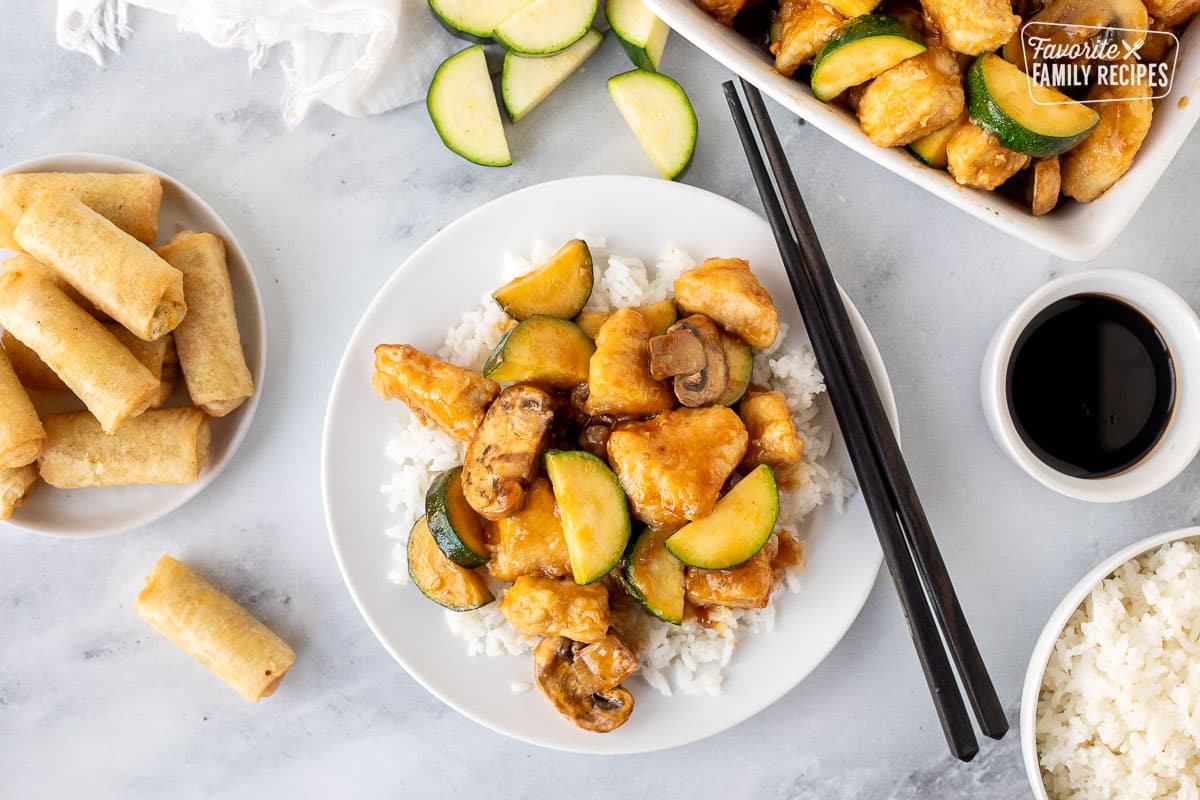 Chopsticks resting on a plate with rice and Panda Express Mushroom Chicken. Spring rolls, rice and soy sauce on the side.