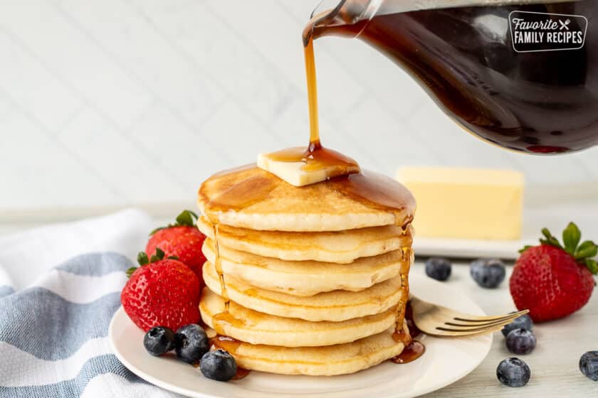 Homemade Maple Syrup pouring over a tall stack of pancakes with butter. Berries on the side.