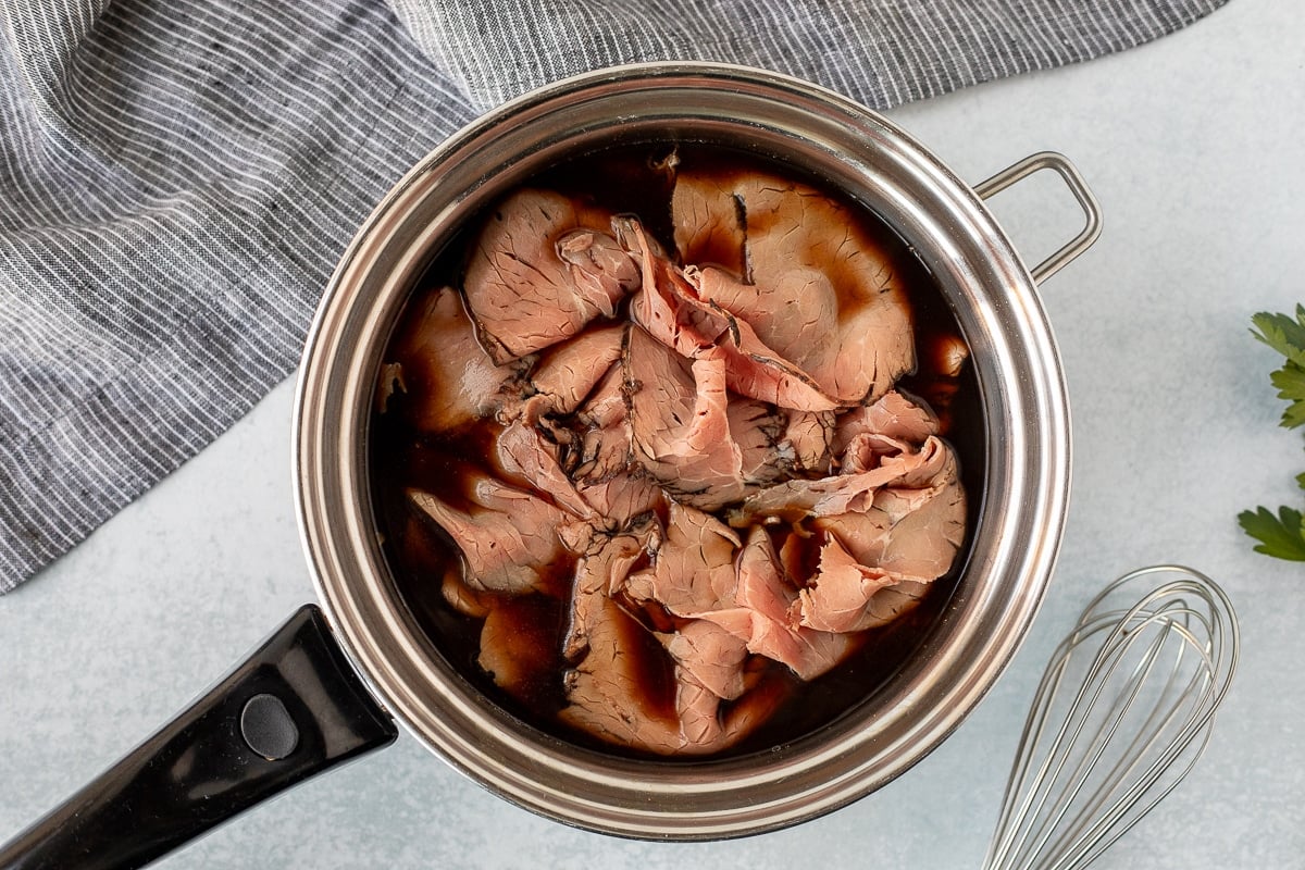 Pan with au jus and sliced roast beef for French Dip Sandwiches.