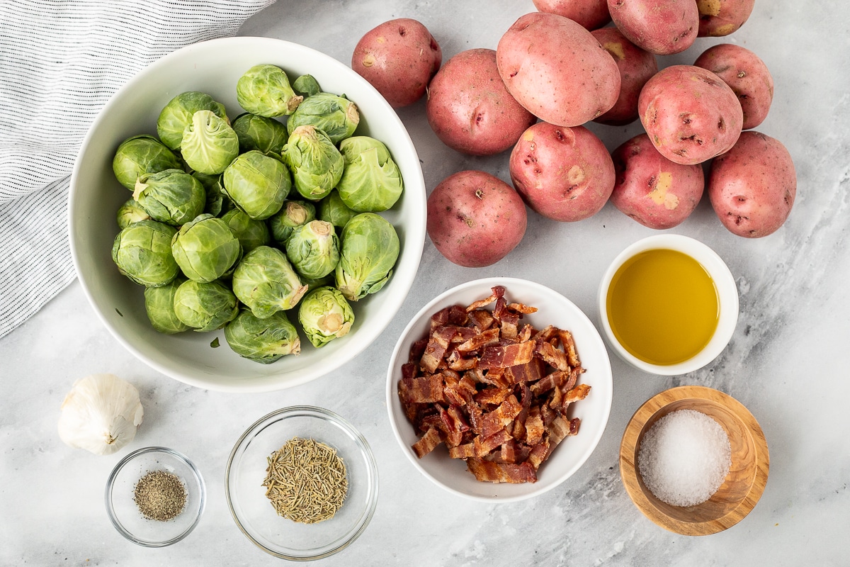 Ingredients to make Roasted Potatoes and Brussels Sprouts including red potatoes, Brussels sprouts, bacon, olive oil, salt, rosemary, pepper and garlic.
