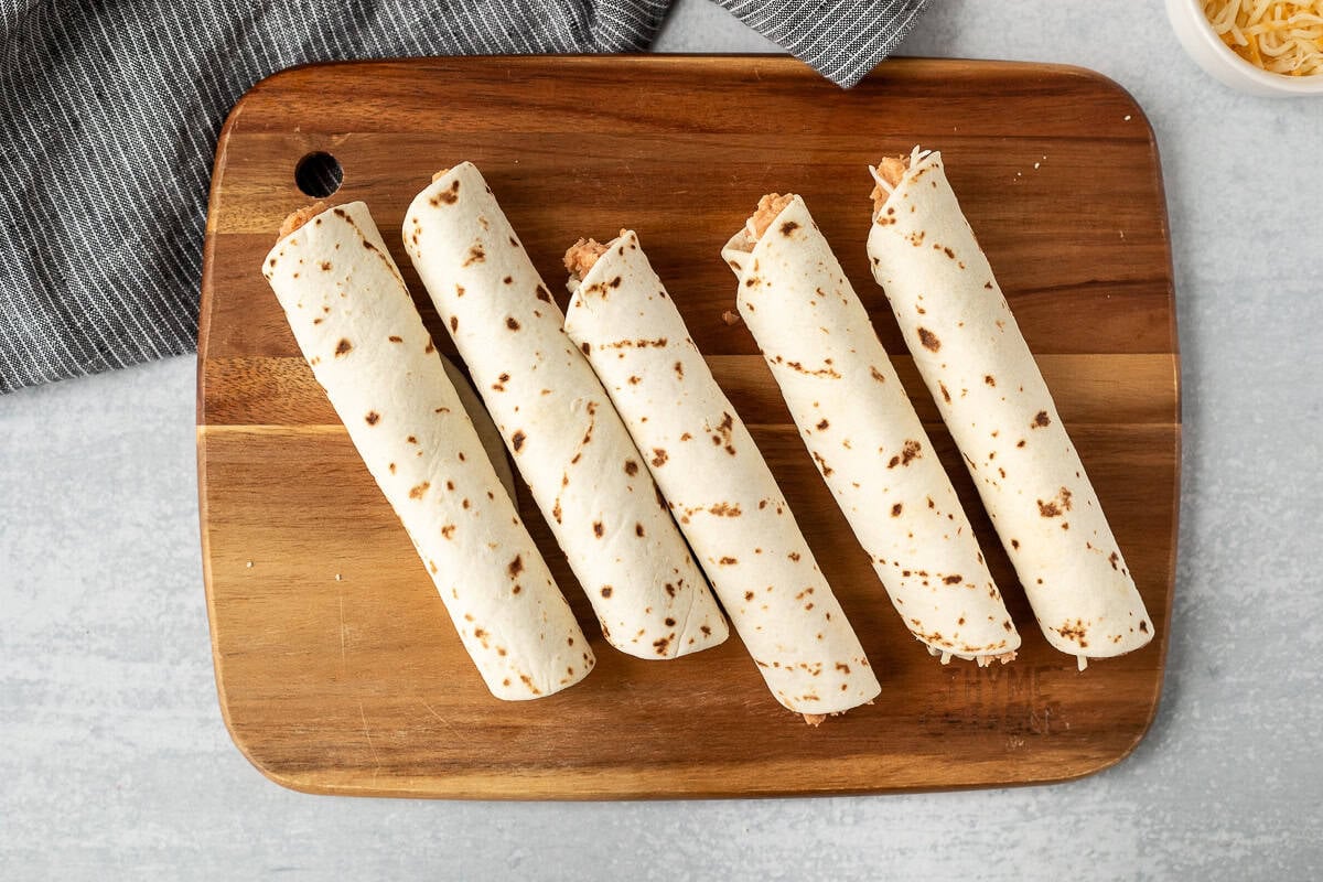 Five rolled bean burritos on a cutting board.