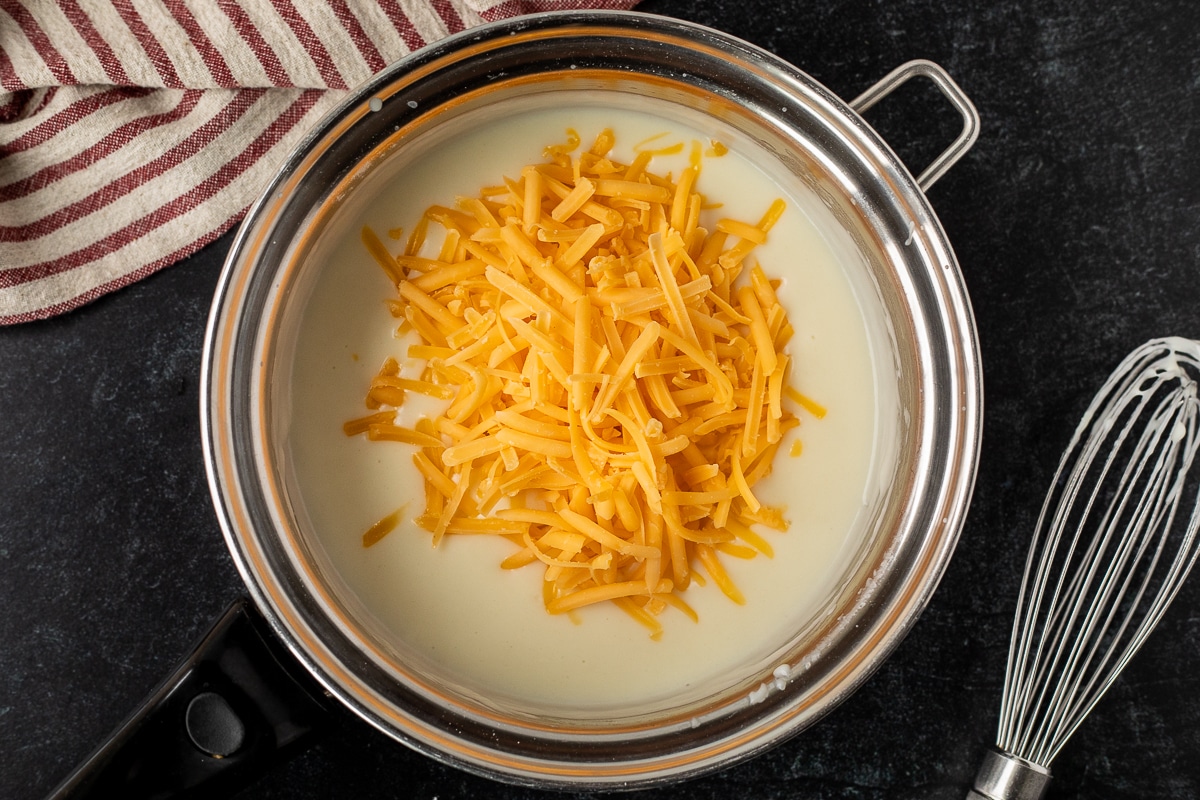 Pan with creamy sauce and shredded cheddar cheese and a whisk on the side.