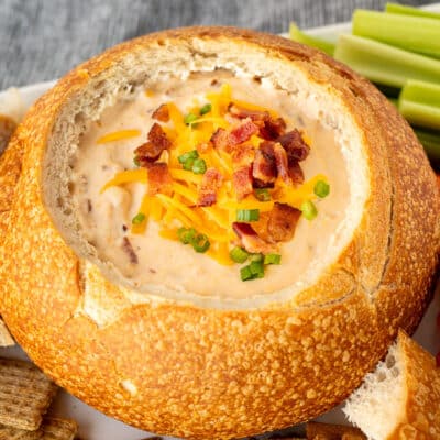 Close up of a bread bowl with warm bacon cheese dip topped with cheddar cheese, bacon and sliced green onions.