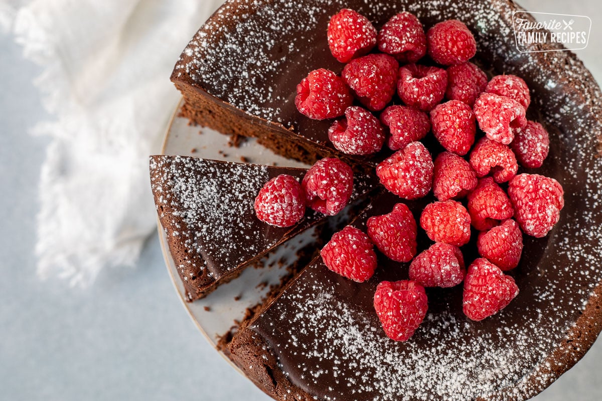 Top view of a Gluten Free Chocolate Cake with a cut out slice. Raspberries and powdered sugar resting on top.