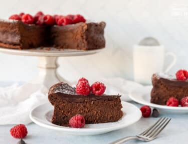 Side view of two slices of Gluten Free Chocolate Cake on plates with fresh raspberries and powdered sugar on top.