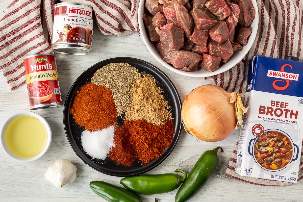 Ingredients to make Texas Chili including beef, beef broth, onion, jalapeños, chipotle peppers, tomato paste, spices, garlic and olive oil.