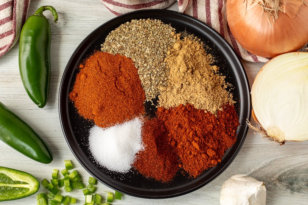 Plate with spices for Texas Chili including chipotle chili powder, chili powder, cumin, oregano, sugar and spoked paprika.