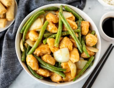 Large bowl of Panda Express String Bean Chicken next to a cup of soy sauce, spring rolls, rice and chop sticks.