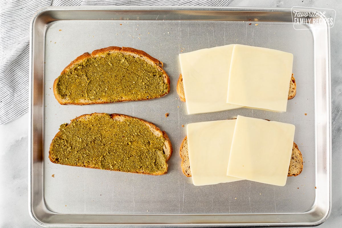 Baking sheet with sliced bread layered with cheese and pesto spread on other half.