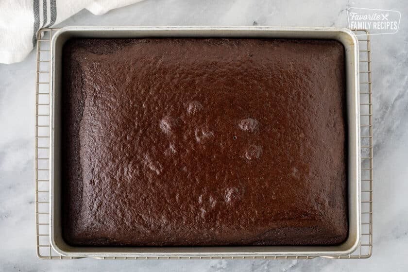 Cake pan with baked chocolate cake cooling on a wire rack.