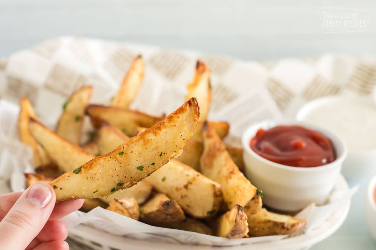 A Baked potato wedge held up in front of a basket full of wedges and ketchup