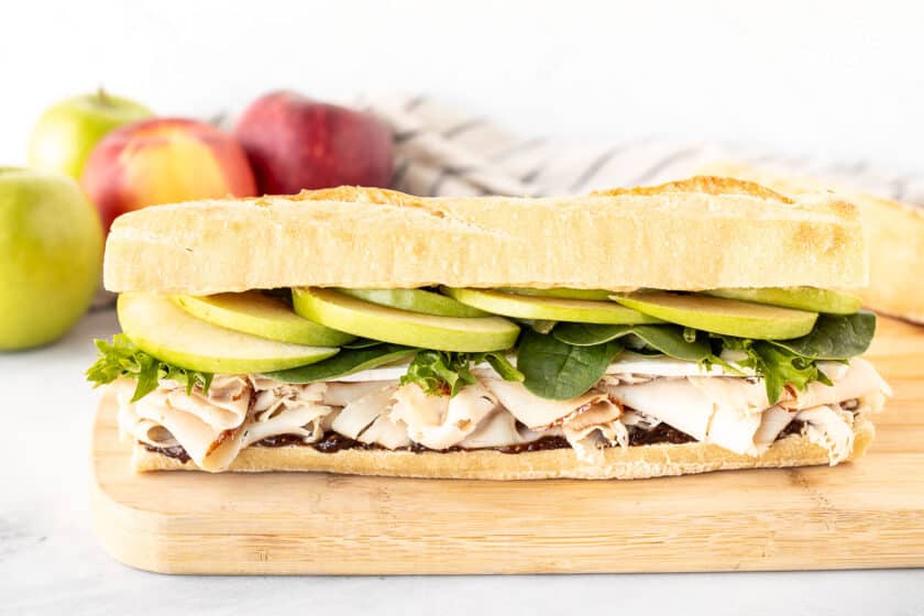 Whole turkey sandwich with brie cheese and apples on a cutting board.