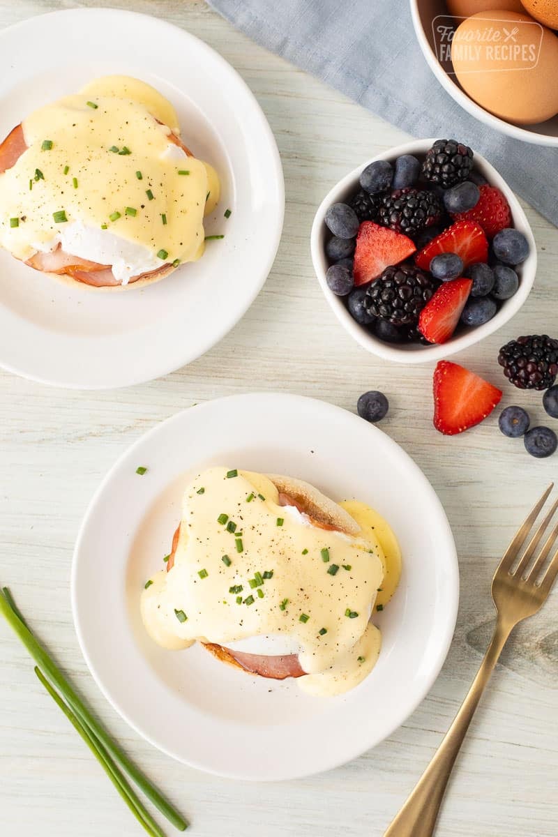 two plates of eggs benedict with a bowl of berries and a bowl of eggs in the background