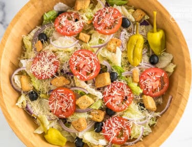 A large bowl with iceberg lettuce, olives, tomatoes, red onions, croutons, pepperoncinis, and parmesan