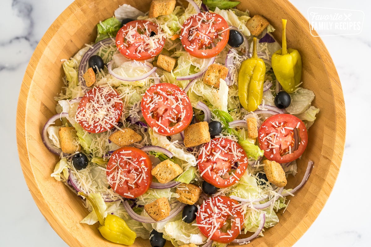 A large bowl with iceberg lettuce, olives, tomatoes, red onions, croutons, pepperoncinis, and parmesan.